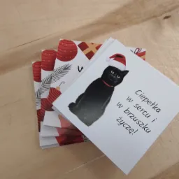 Stos świątecznych kartek z życzeniami z czarnym kotem w czapce Mikołaja, wykonanych w Warszawie, leżących na drewnianym blacie.