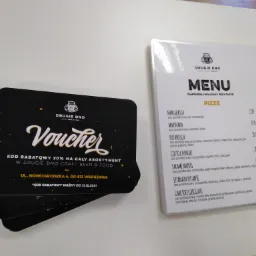Stos czarnych voucherów rabatowych z białym napisem 'Voucher' i logo 'Drugie Dno', leżący obok menu z pizzami restauracji 'Drugie Dno'.