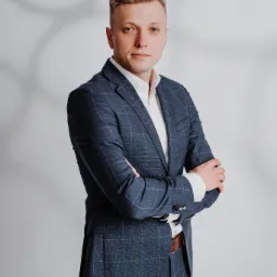 Portret mężczyzny w garniturze w kratę, z założonymi rękami, na jasnym tle z cieniem. Profesjonalny wizerunek prawnika lub doradcy.