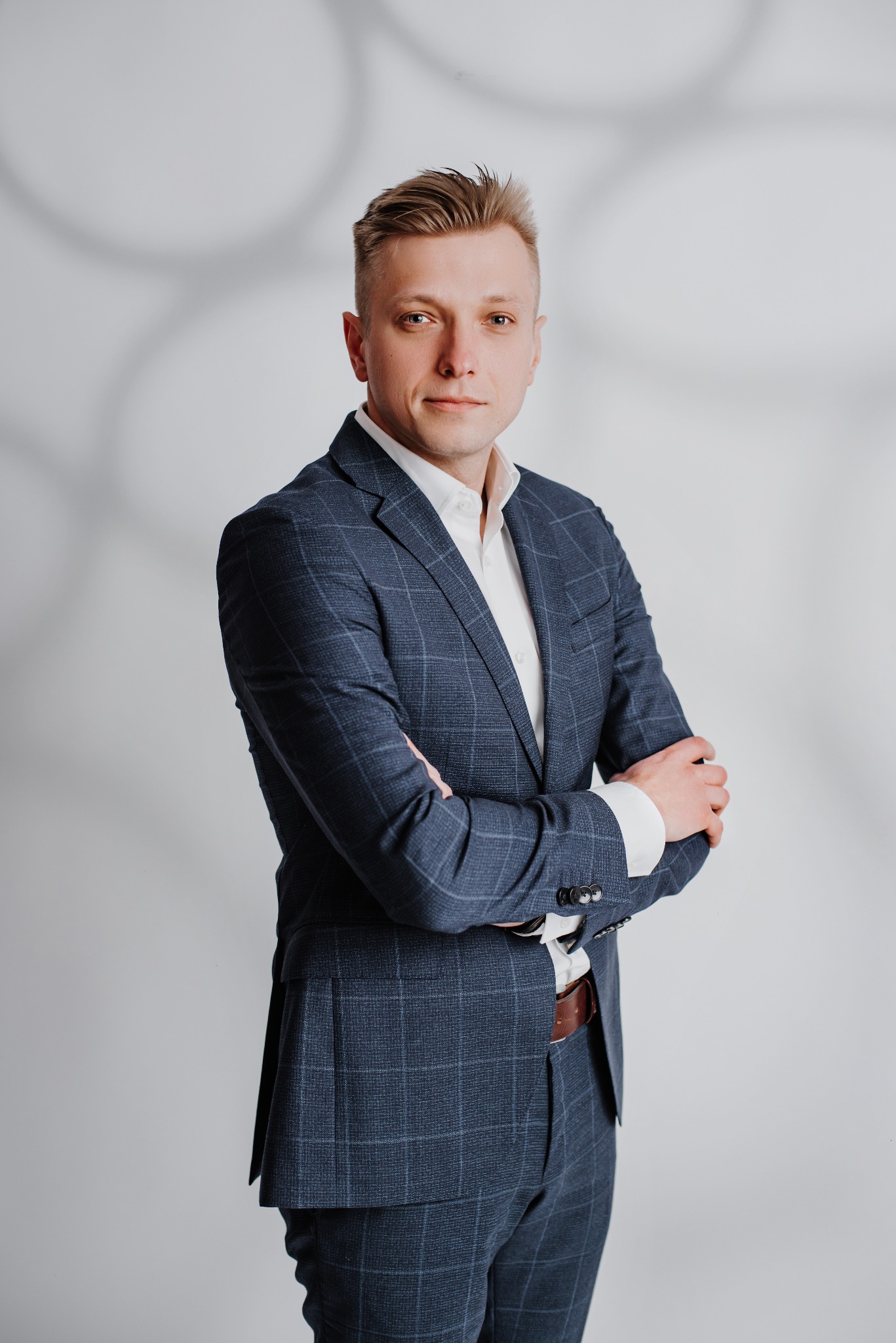 Portret mężczyzny w garniturze w kratę, z założonymi rękami, na jasnym tle z cieniem. Profesjonalny wizerunek prawnika lub doradcy.