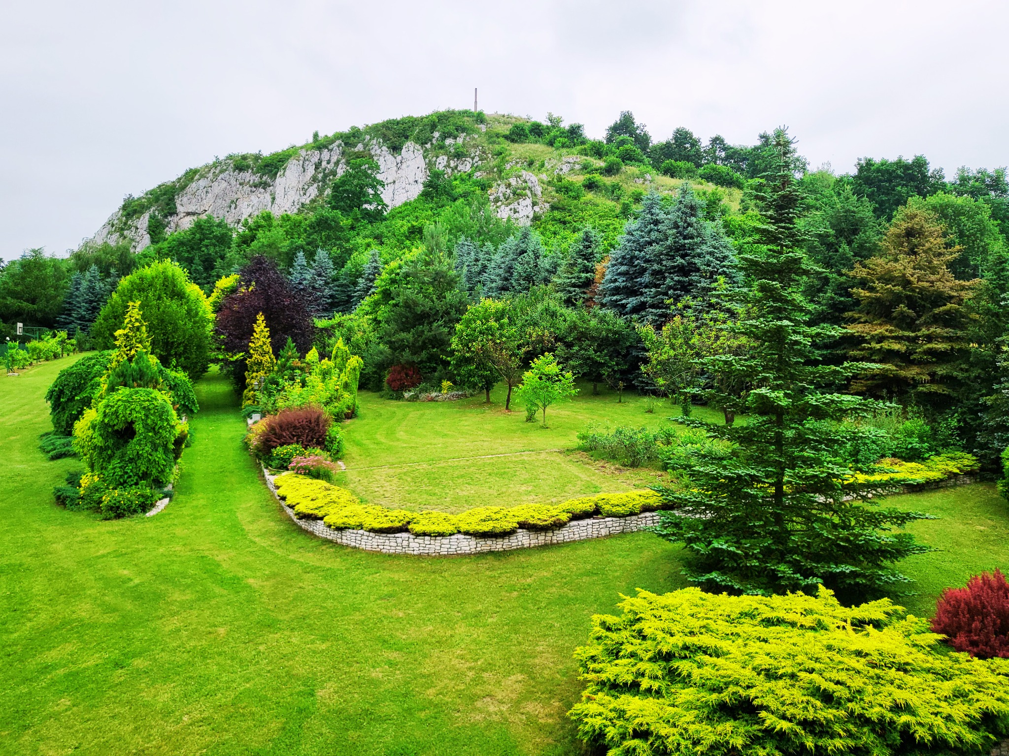 Widok na zadbany ogród z różnorodną roślinnością, w tle skalista góra z pomnikiem na szczycie.