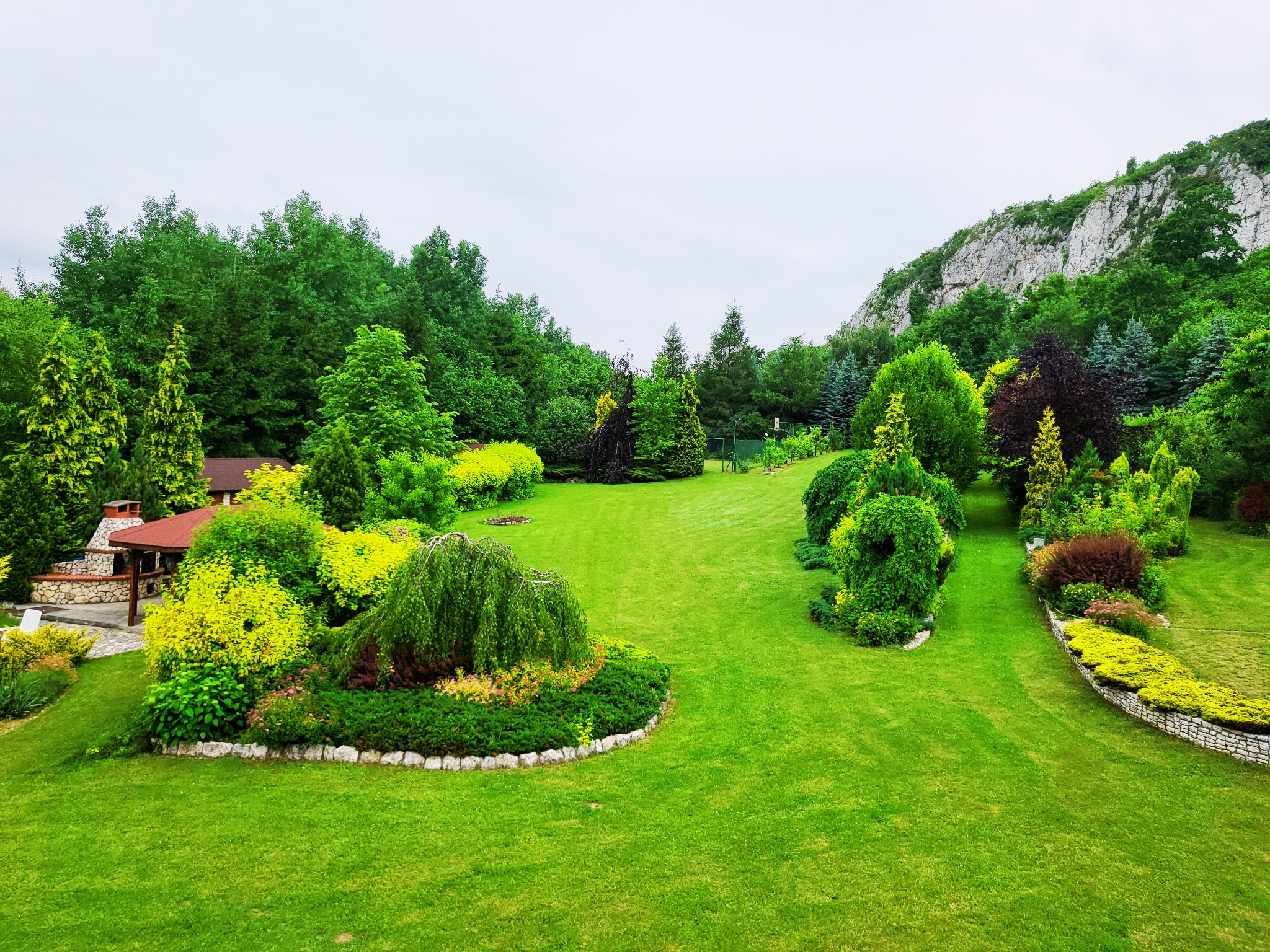 Rozległy, zadbany ogród w Jerzmanowicach z równo przystrzyżonym trawnikiem, ozdobnymi krzewami i drzewami, w tle widoczna skała i budynek z kamiennym grillem.