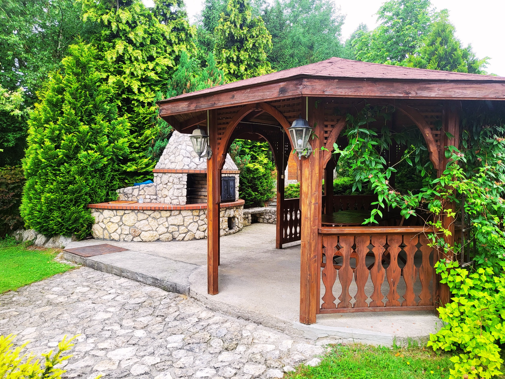 Drewniana altana z grillem murowanym z kamienia, otoczona zielenią drzew i krzewów.