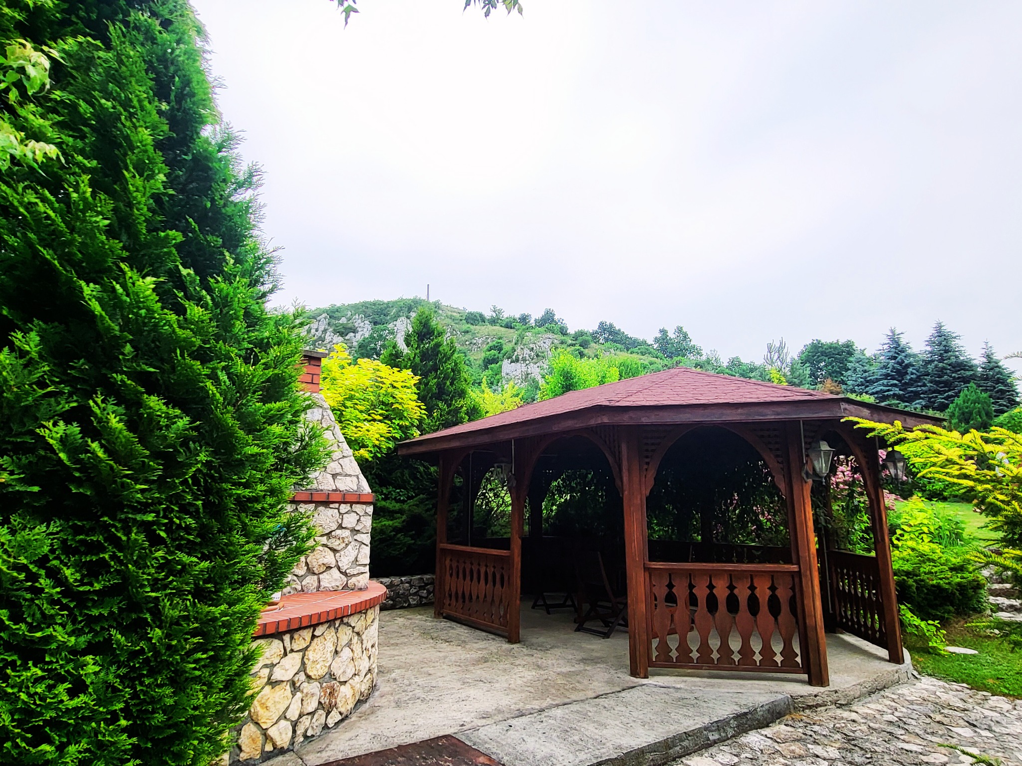 Drewniana altana z czerwonym dachem, obok kamienny grill, otoczona zielenią i drzewami, w tle wzgórze.