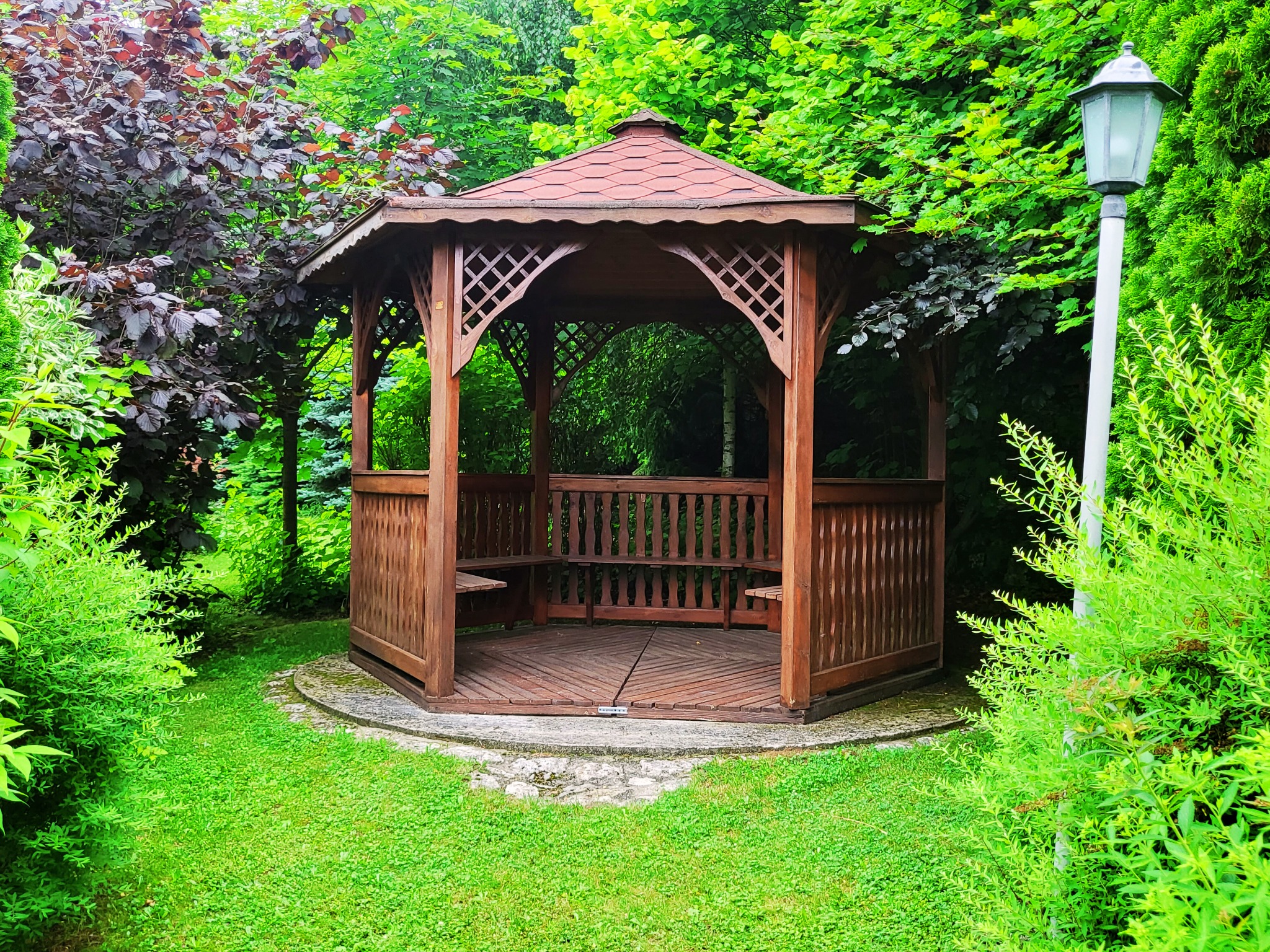 Drewniana altana ogrodowa z czerwonym dachem, otoczona bujną zielenią i oświetlona latarnią, widoczna z poziomu trawnika.