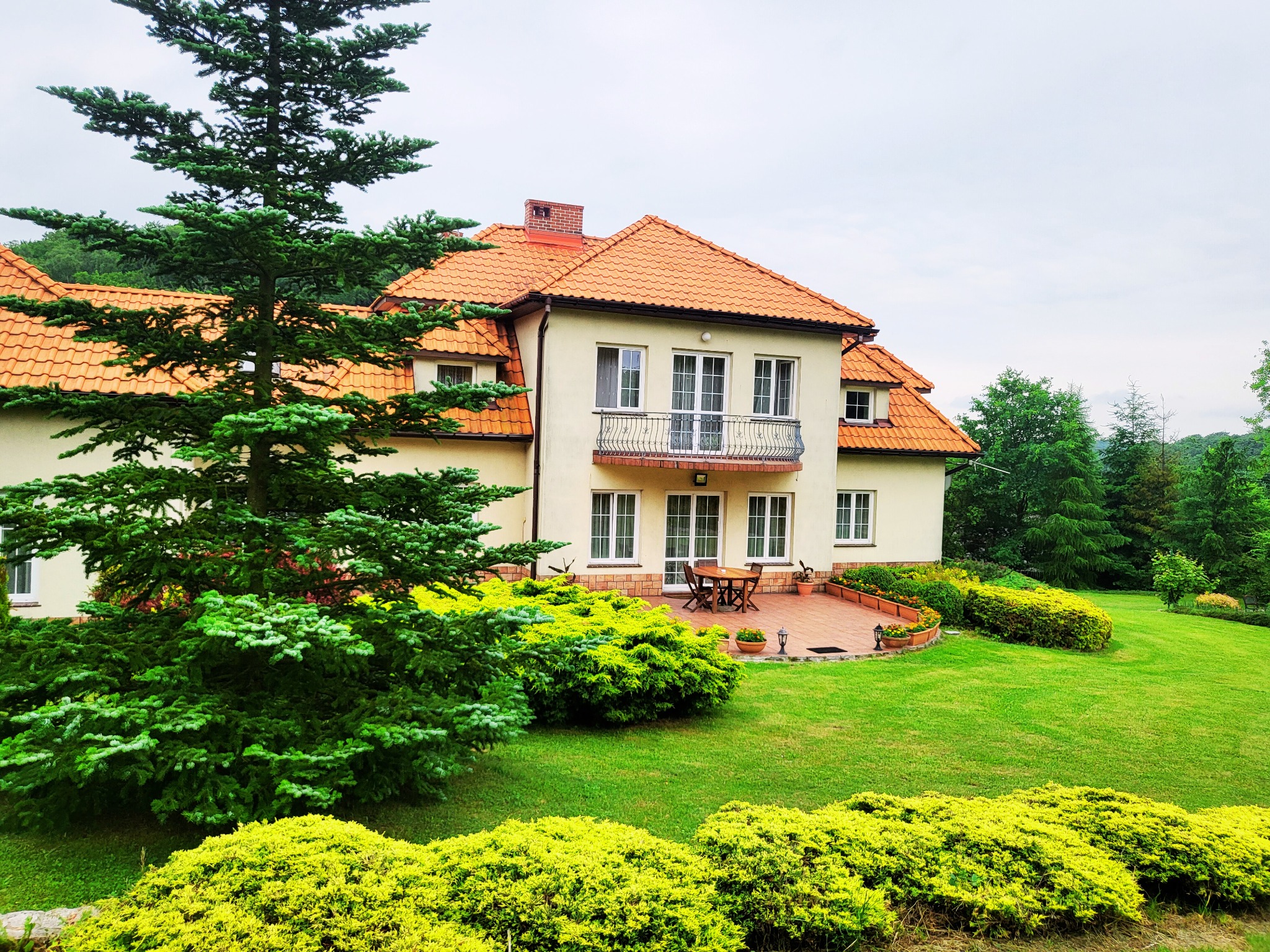 Dwupiętrowy dom z beżową elewacją i pomarańczową dachówką, otoczony zielonym trawnikiem i krzewami, z widocznym tarasem i balkonem.