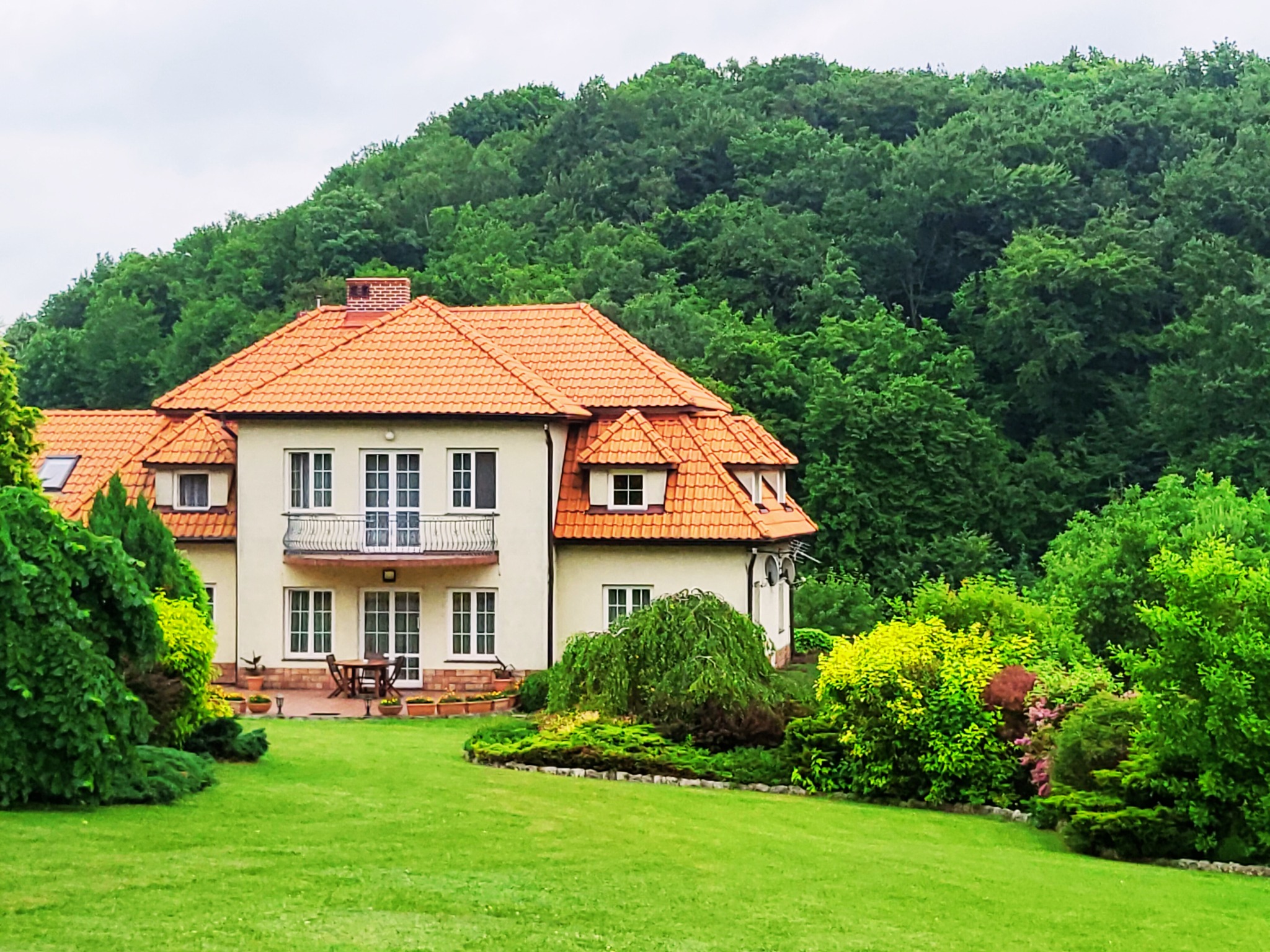 Dwupiętrowy dom z pomarańczową dachówką, balkonem i tarasem, otoczony zielonym trawnikiem i drzewami.