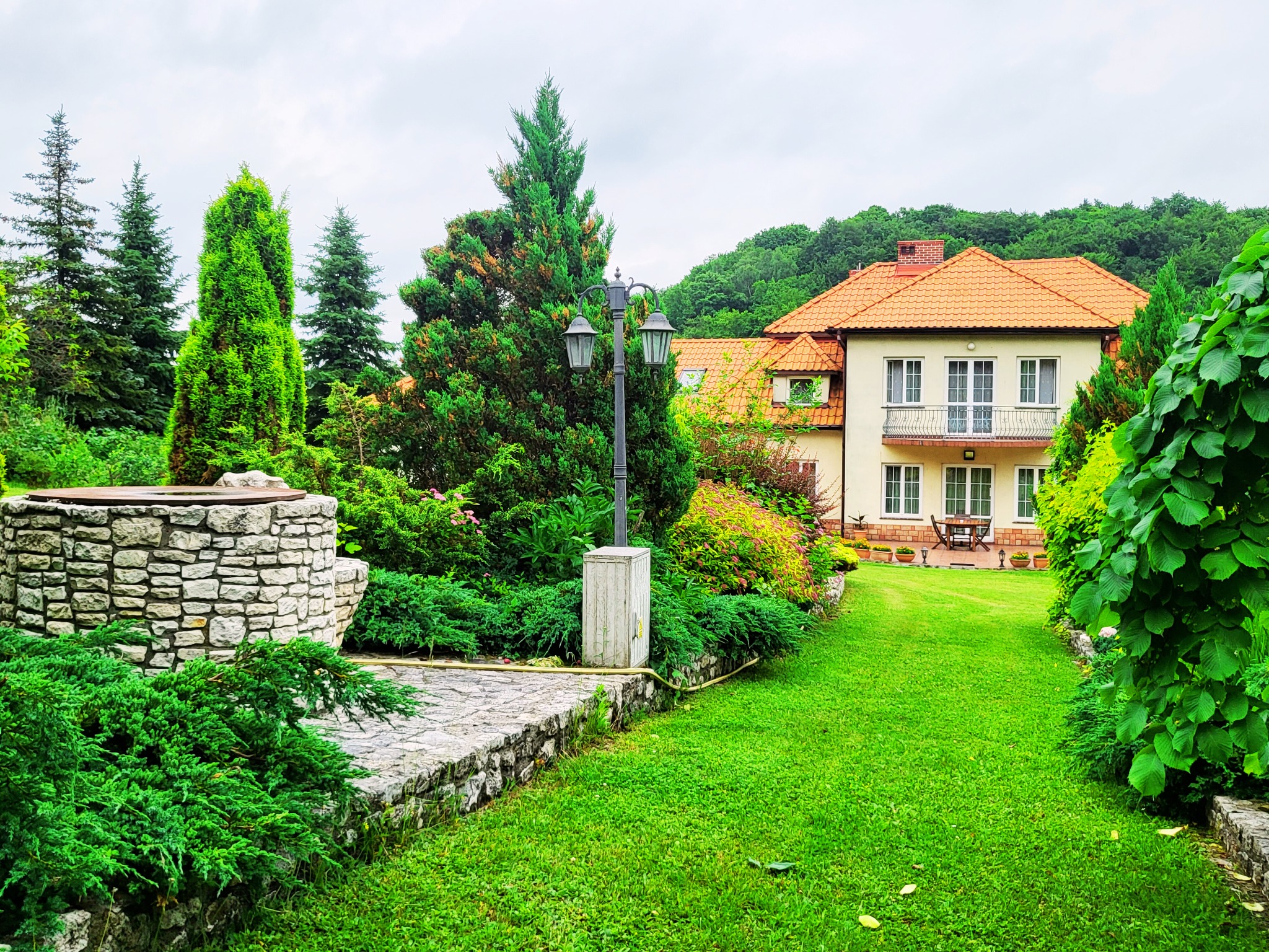 Dwupiętrowy dom z pomarańczowym dachem, widoczny z zadbanego ogrodu z kamienną studnią i ścieżką.