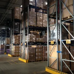 Wnętrze magazynu z wysokimi regałami wypełnionymi kartonami na paletach, pracownik w odblaskowej kamizelce przechodzi w tle