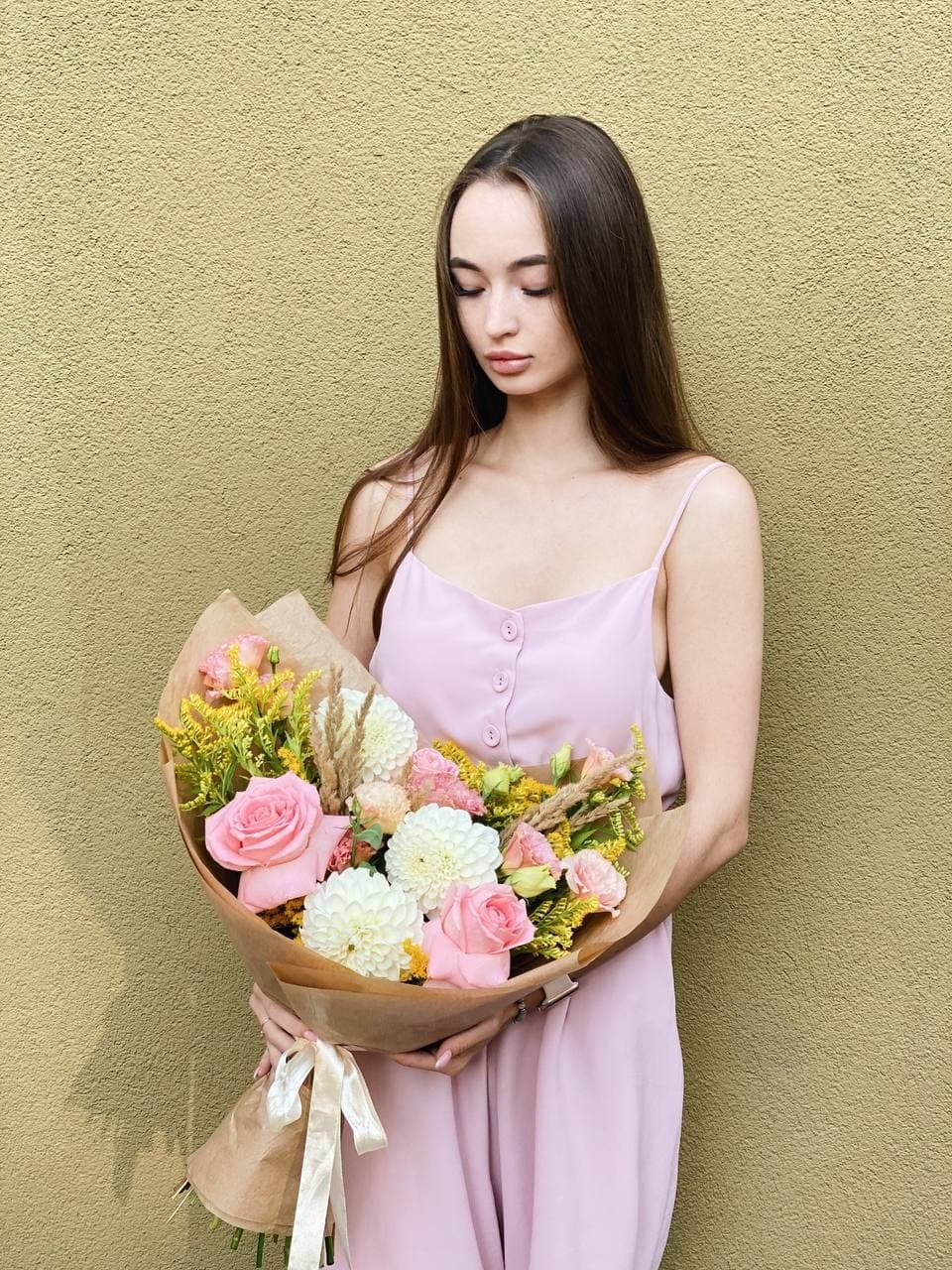 Młoda kobieta w jasnoróżowej sukience trzyma bukiet różowych róż, białych dalii i żółtych kwiatów w brązowym papierze z kremową wstążką na tle teksturowanej oliwkowej ściany.