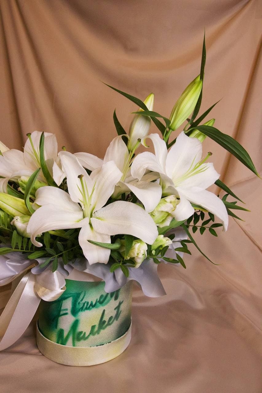 Elegancki bukiet białych lilii i zielonych dodatków w okrągłym, zielono-białym pudełku z napisem 'Flower Market', ozdobiony szarą wstążką na tle beżowej tkaniny.