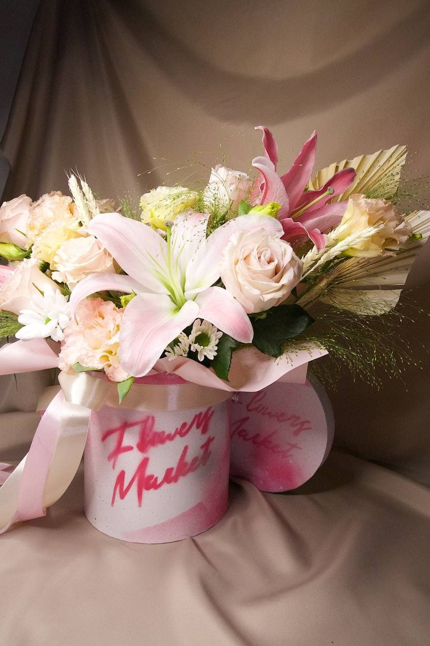 Delikatny bukiet róż, lilii i traw w okrągłym, białym pudełku z napisem 'Flower Market', przewiązany satynową wstążką, na tle beżowej tkaniny.