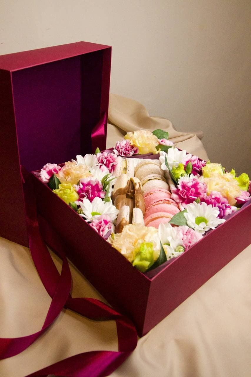 Burgundowy otwarty kartonowy box wypełniony makaronikami, czekoladkami i świeżymi goździkami w odcieniach bieli, różu i żółci, z bordową wstążką na beżowym tle.