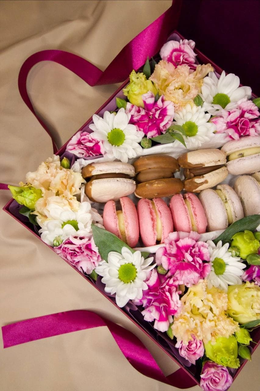 Elegancki kosz prezentowy z Lublina wypełniony kolorowymi makaronikami i świeżymi kwiatami, ozdobiony fuksjową wstążką w kształcie serca, na beżowym tle.