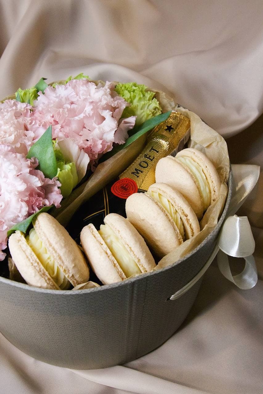 Okrągły, szary kosz prezentowy z beżowymi makaronikami, butelką szampana Moët & Chandon i bukietem różowych kwiatów na beżowym tle.