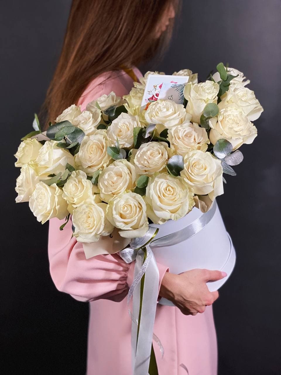 Kobieta w różowej sukience trzyma biały okrągły kosz z kremowymi różami, eukaliptusem i kartką z życzeniami, przewiązany srebrną wstążką, na ciemnym tle.