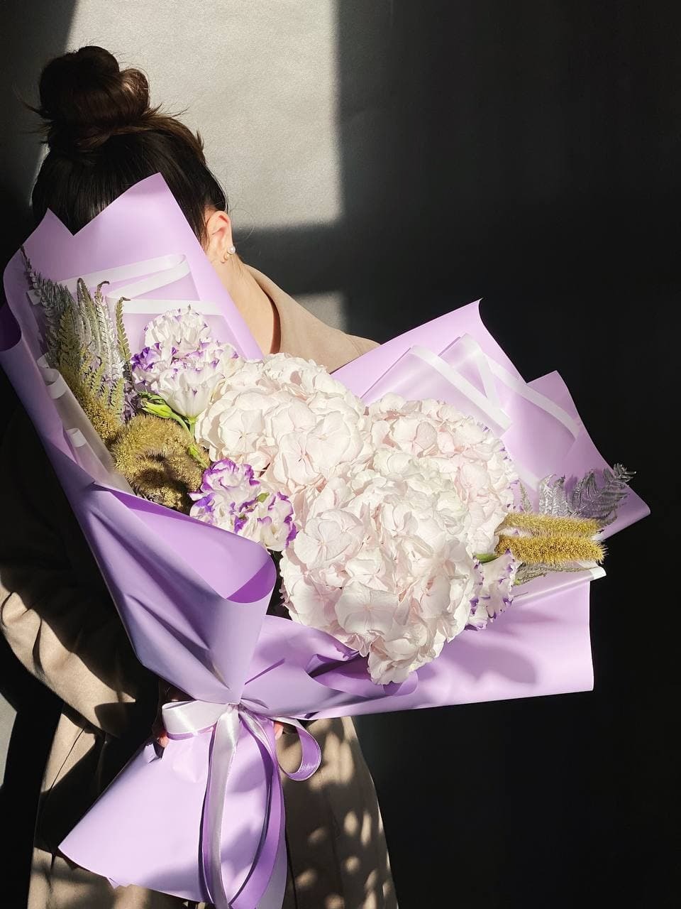 Kobieta trzyma bukiet białych hortensji i fioletowych kwiatów w liliowym papierze z białą wstążką, oświetlony bocznym światłem.