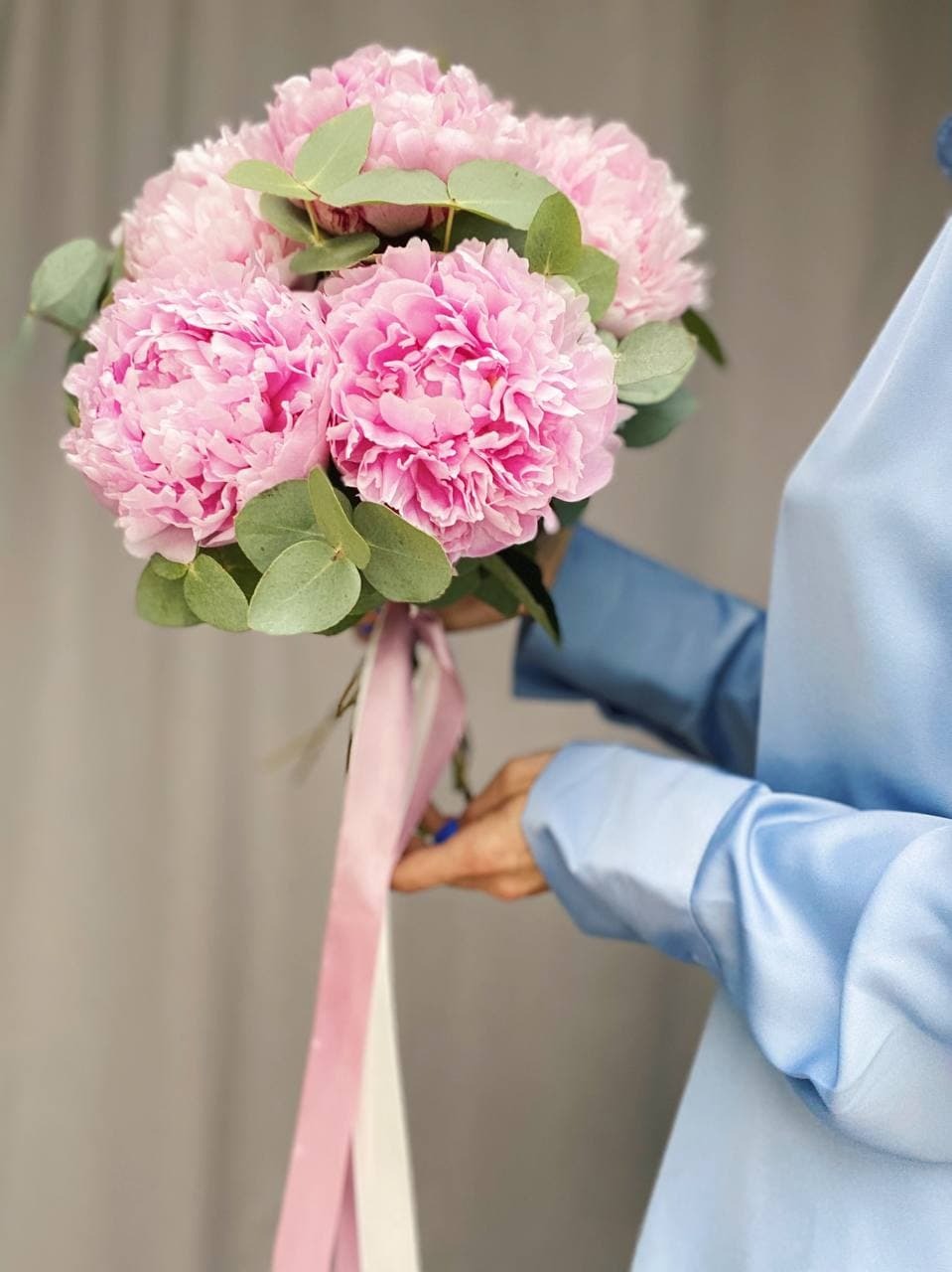Delikatny bukiet z różowych piwonii i eukaliptusa, przewiązany różową i białą wstążką, trzymany przez osobę w błękitnej koszuli.