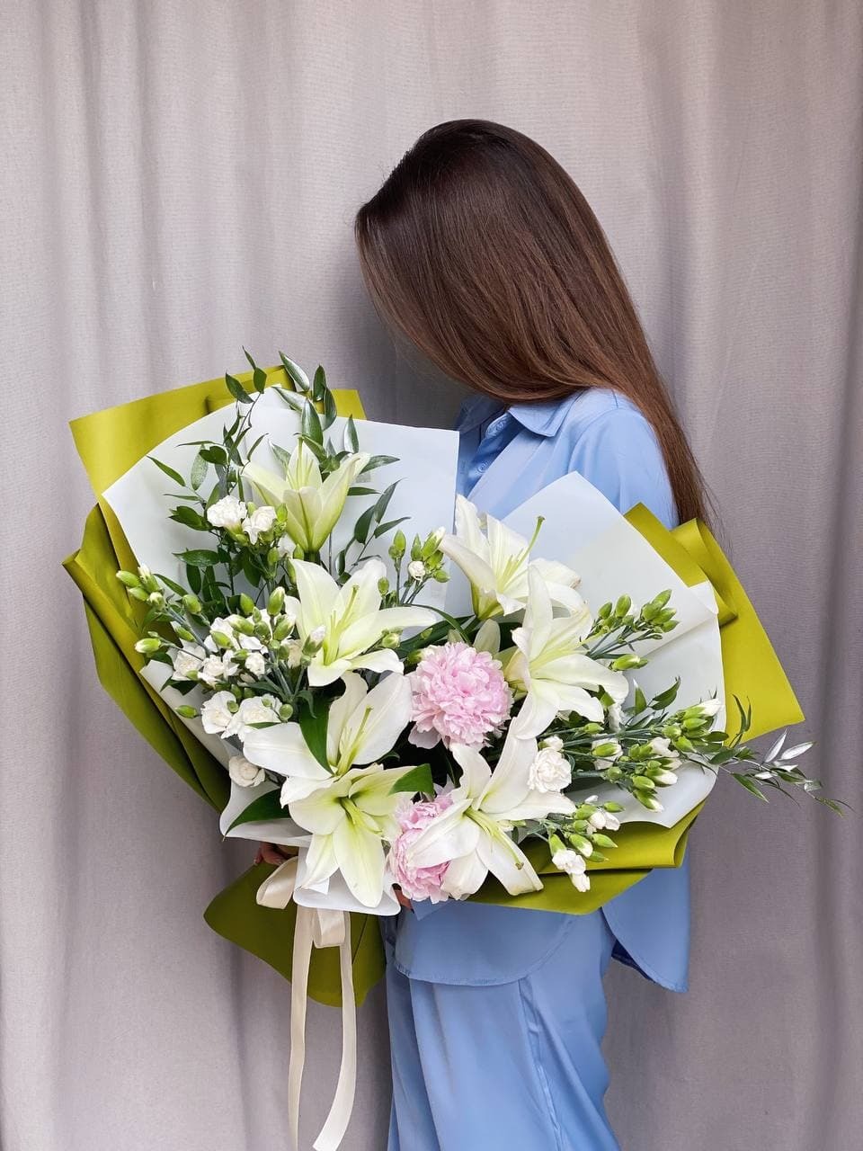 Kobieta w niebieskiej koszuli trzyma duży bukiet białych lilii i różowych goździków zawinięty w zielony i biały papier z kremową wstążką.