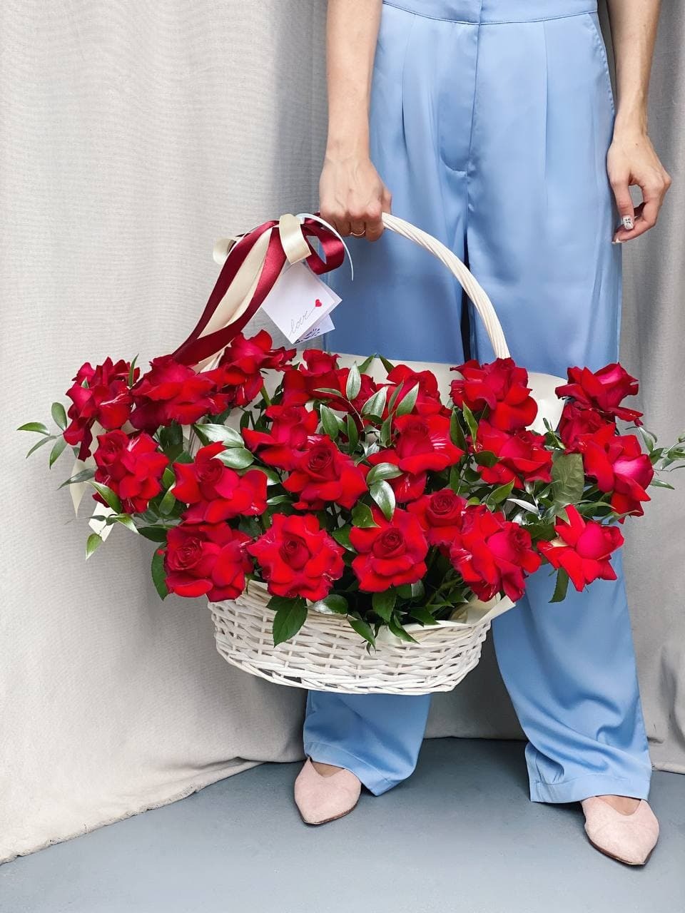 Osoba w błękitnych spodniach trzyma biały wiklinowy kosz wypełniony czerwonymi różami, ozdobiony wstążkami i kartką z napisem 'love'.