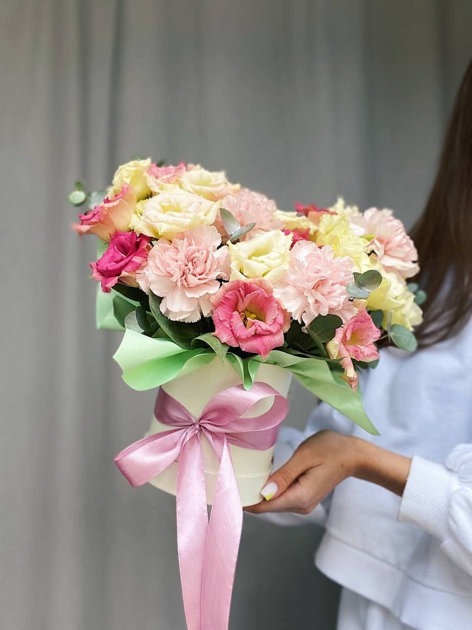 Kobieta trzyma biały pudełko z mieszanką różowych, kremowych i żółtych kwiatów, przewiązane satynową, różową wstążką na tle szarej zasłony.