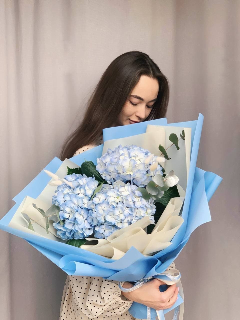 Kobieta z długimi włosami trzyma duży bukiet błękitnych hortensji w błękitnym i kremowym papierze, przewiązany srebrną wstążką, z delikatnym uśmiechem.