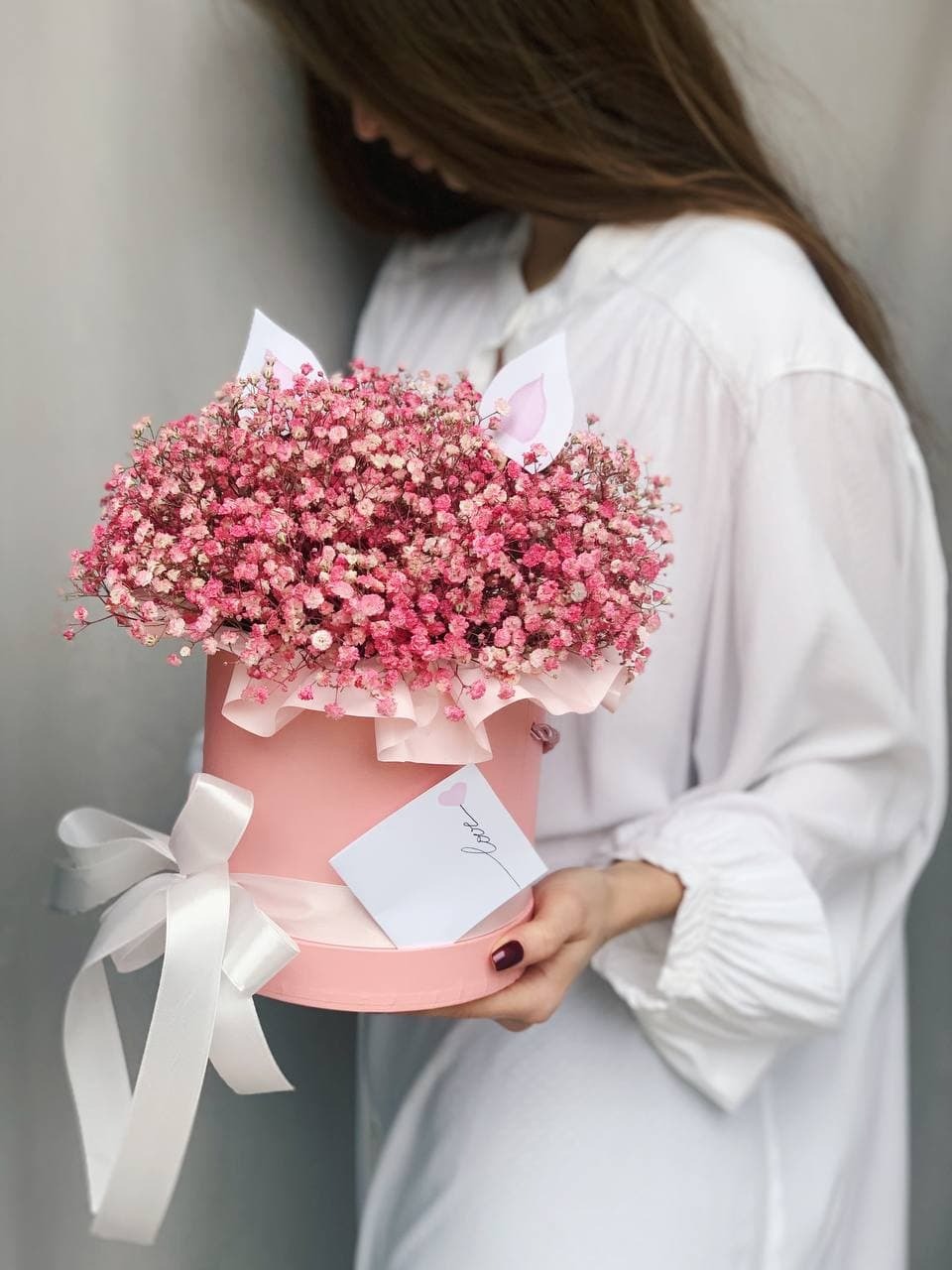 Kobieta trzyma różowy okrągły pudełko z bukietem różowych kwiatów gipsówki, ozdobione białymi uszkami królika i kartką z napisem 'love' oraz białą wstążką, na tle szarej ściany.