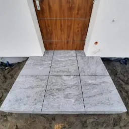 Ułożony z jasnoszarych płyt kamiennych podest przed wejściem do domu z brązowymi drzwiami, widoczne ślady ziemi po bokach.