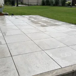 Świeżo ułożony taras z jasnoszarych, kwadratowych płyt betonowych, z widocznymi śladami wody po myciu, przylegający do budynku z trawnikiem i ozdobnymi krzewami w tle.