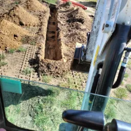 Wykopany wąski rów w ziemi widziany z kabiny minikoparki, hałdy ziemi obok wykopu, w tle budynek mieszkalny i pomarańczowe rury.