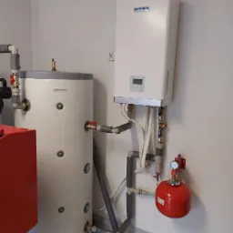 Zamontowana pompa ciepła marki Sevra wraz z zasobnikiem Totenso i czerwonym zbiornikiem wyrównawczym w pomieszczeniu w miejscowości Rusiec, widoczne rury i przyłącza.