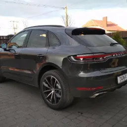 Ciemnoszary Porsche Macan, widziany z tyłu pod kątem, z tablicą rejestracyjną 'DARKFOL', stojący na kostce brukowej przed ogrodzeniem.