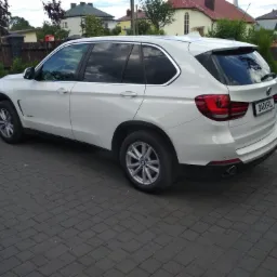 Białe BMW X5 z przyciemnionymi szybami zaparkowane na brukowanym podjeździe przed domem z brązowym ogrodzeniem i zielenią w tle. Tablica rejestracyjna z napisem 'DARKFUL'.