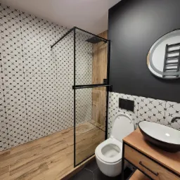 Nowoczesna łazienka z kabiną prysznicową typu walk-in, drewnopodobnymi płytkami na podłodze i ścianie w kabinie, czarno-białymi płytkami wzorzystymi na ścianach, czarną ścianą, owalnym lustrem...