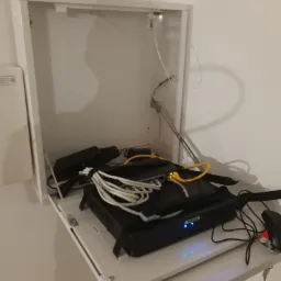Router z podłączonymi kablami sieciowymi umieszczony w białej szafce instalacyjnej z opuszczanym blatem.