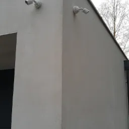 Dwie białe kamery monitoringu zamontowane na rogu szarego budynku, widok z dołu na tle bezlistnego drzewa.