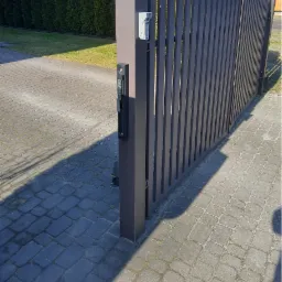 Nowoczesna, grafitowa brama z pionowych sztachet z zamontowanym wideodomofonem i elektrozaczepem, na tle kostki brukowej i żywopłotu.