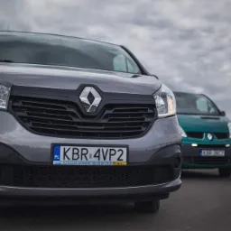 Dwa busy Renault Trafic zaparkowane na zewnątrz; szary na pierwszym planie z włączonym światłem mijania, zielony w tle, pochmurne niebo.