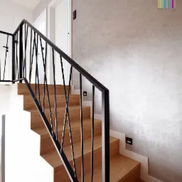 Drewniane schody z czarną, geometryczną balustradą na tle ściany z efektem betonu i białych drzwi.