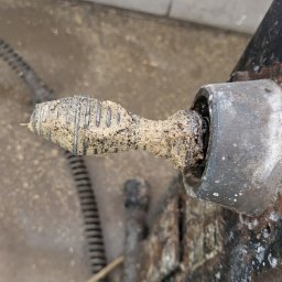 KAN-ŁUK USŁUGI KANALIZACYJNE - Kret do rur, pokryty zaschniętym brudem i resztkami po czyszczeniu, umieszczony w uchwycie wiertarki. Widoczne ślady korozji na metalowych elementach i zabrudzenia na podłożu.