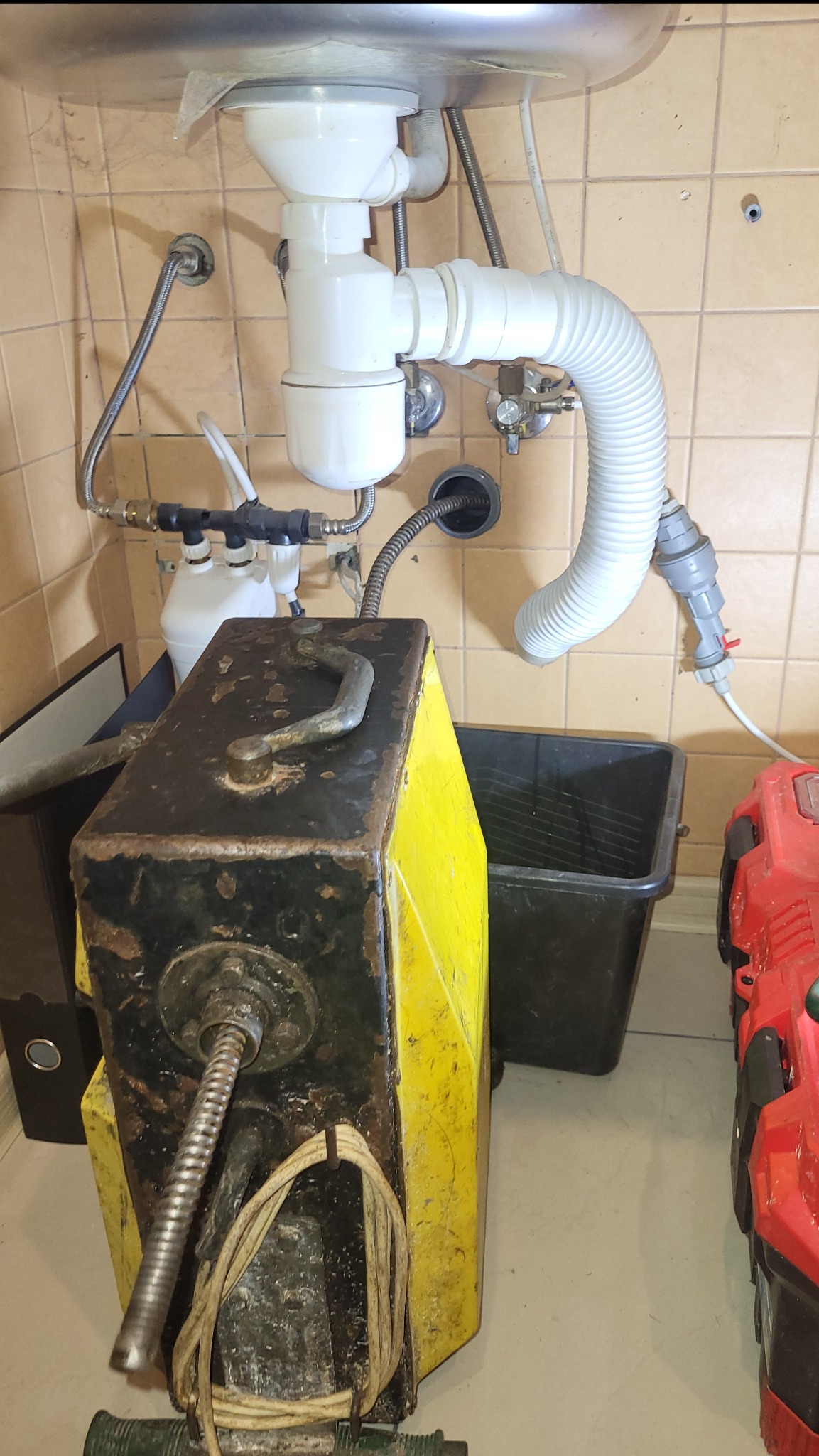 Metalowa spirala wchodzi do otworu kanalizacyjnego pod zlewem. Obok stoi żółto-czarna maszyna do przepychania rur i czarny pojemnik.