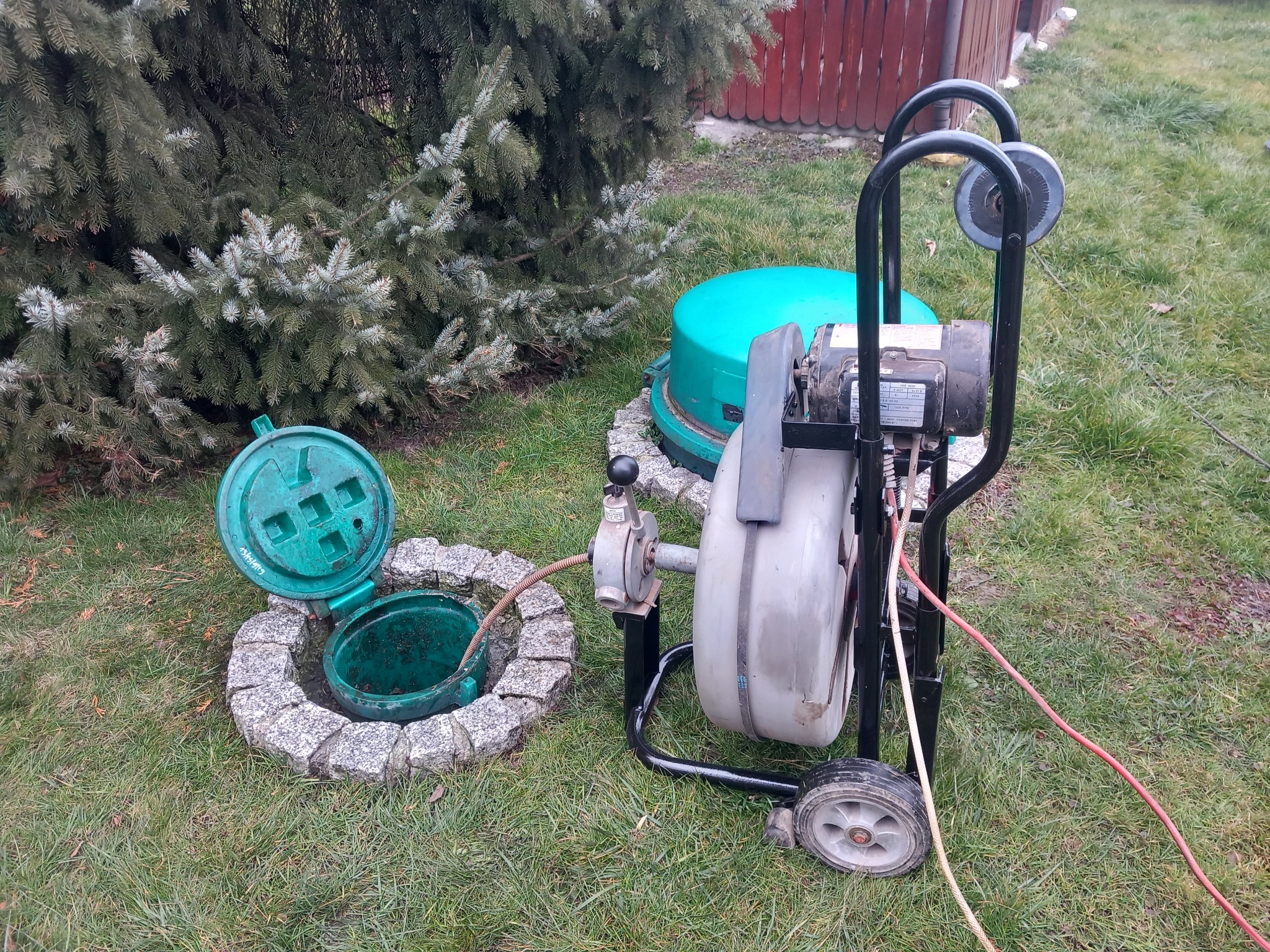 Profesjonalna maszyna do czyszczenia kanalizacji obok otwartego, zielonego włazu kanalizacyjnego otoczonego kamieniami, na trawniku.