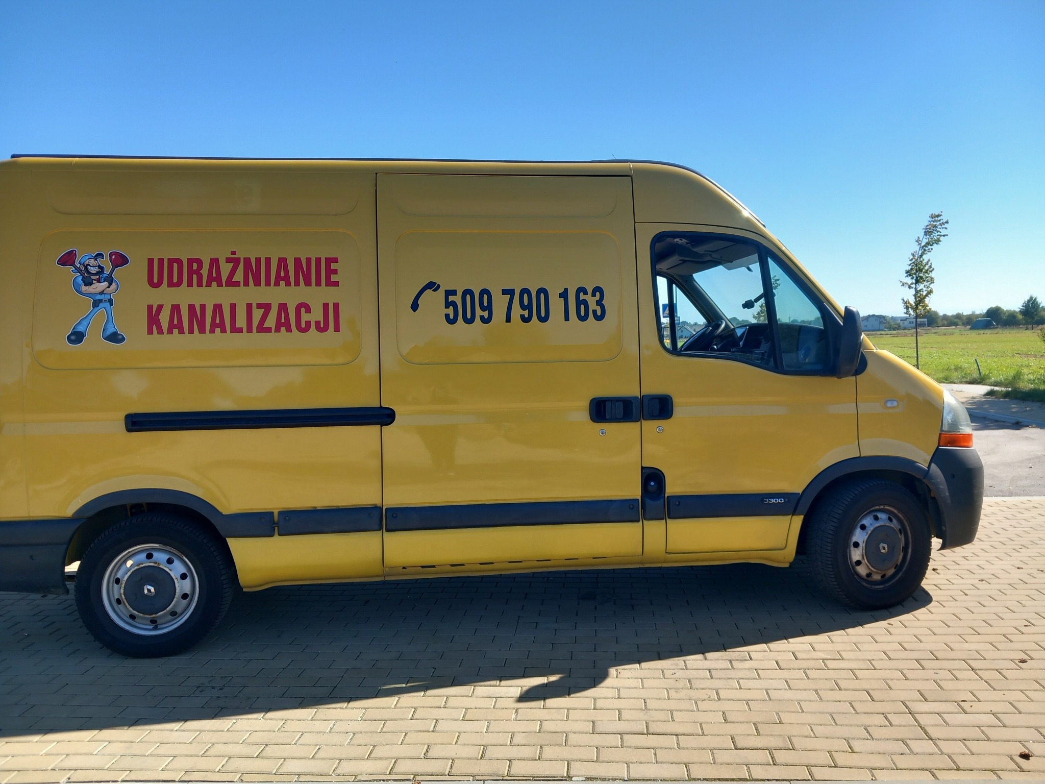 Żółty samochód dostawczy z nadrukowanym numerem telefonu i postacią hydraulika trzymającego przepychacze, zaparkowany na kostce brukowej w słoneczny dzień.
