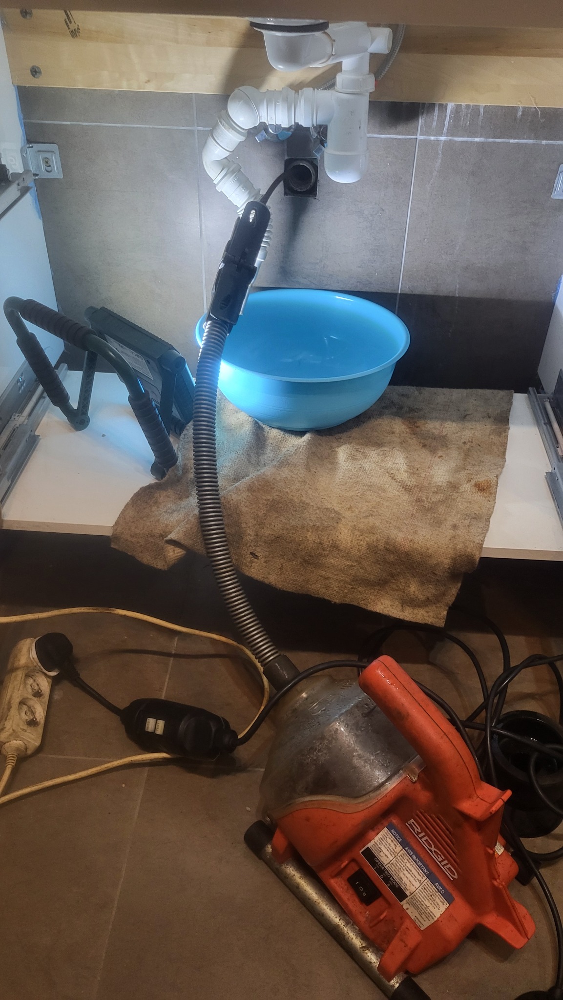 Pomarańczowa elektryczna przepychaczka RIDGID z wężem wprowadzanym do odpływu umywalki, pod którym stoi niebieska miska na brudnej szmacie, widoczne też gniazdko elektryczne i przedłużacz.