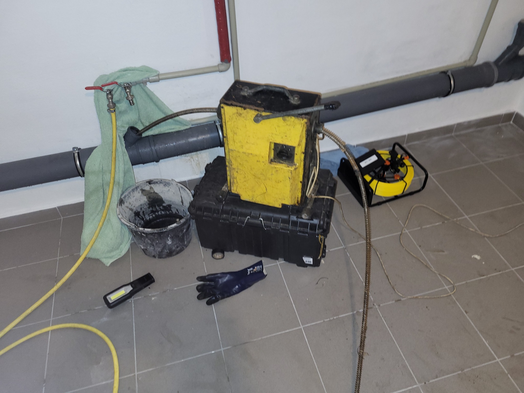 Żółto-czarne urządzenie do czyszczenia rur ustawione na czarnej skrzynce, obok wiadro z brudną wodą, rękawice ochronne i lampa warsztatowa na kafelkach podłogowych, w tle rura kanalizacyjna...