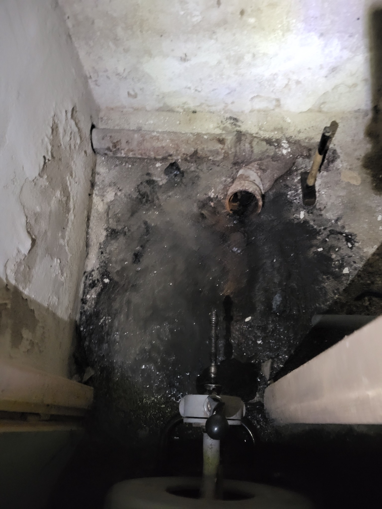 Zalana, brudna podłoga piwnicy z wystającą rurą kanalizacyjną i narzędziem do czyszczenia, widok z góry. Wilgoć na ścianach, ciemne otoczenie.