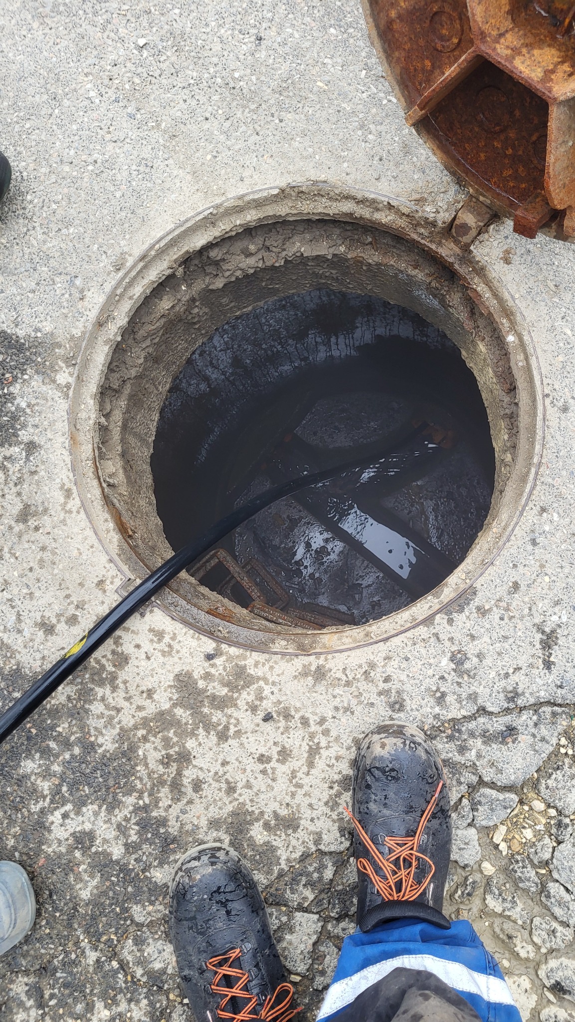 Otwarty właz kanalizacyjny z widocznym kablem do udrażniania i fragmentem drabiny wewnątrz. Obok włazu widoczne buty robocze.