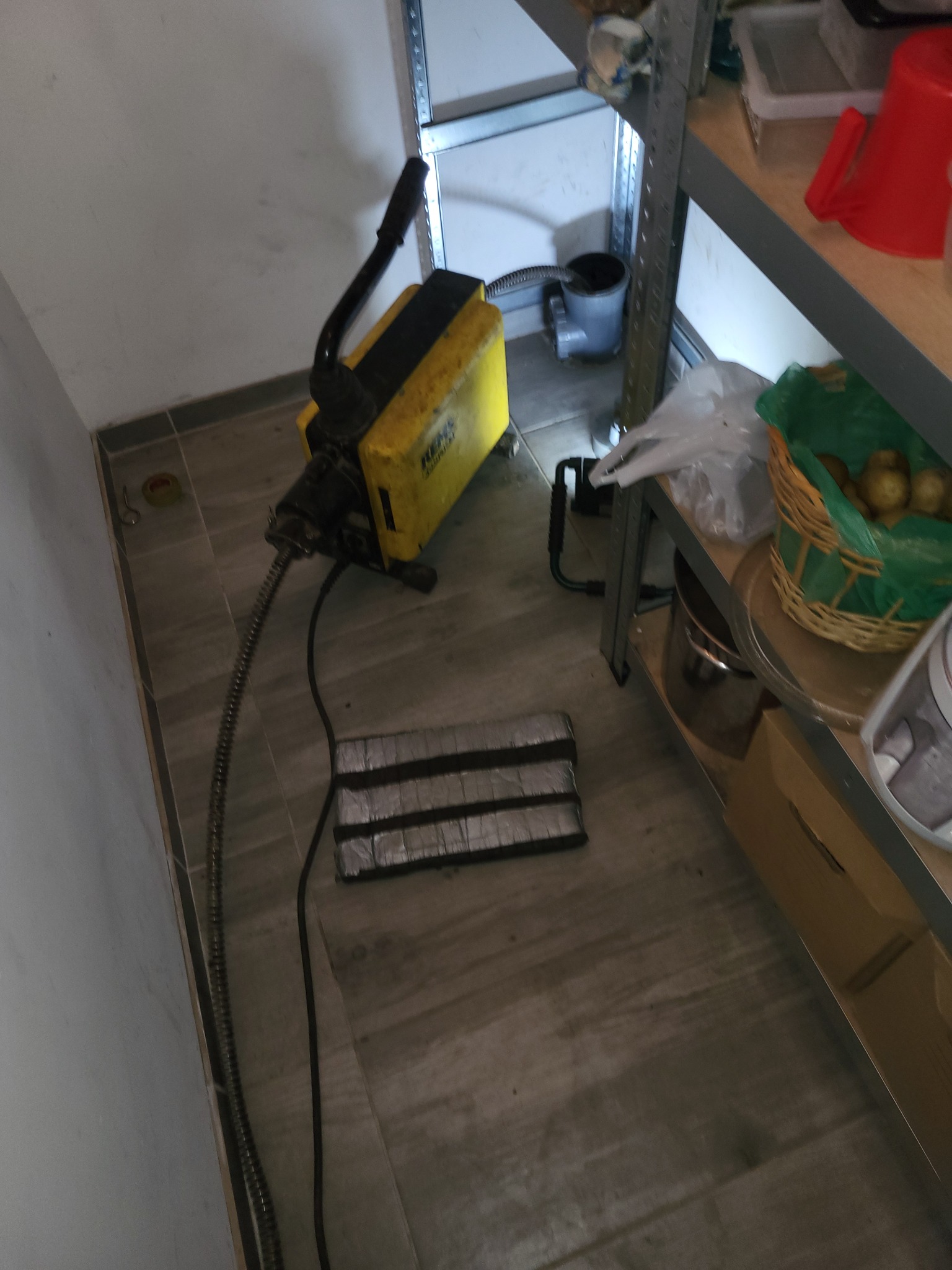 Żółta maszyna do udrażniania rur Rems z wężem na szarej podłodze. W tle metalowy regał z różnymi przedmiotami.