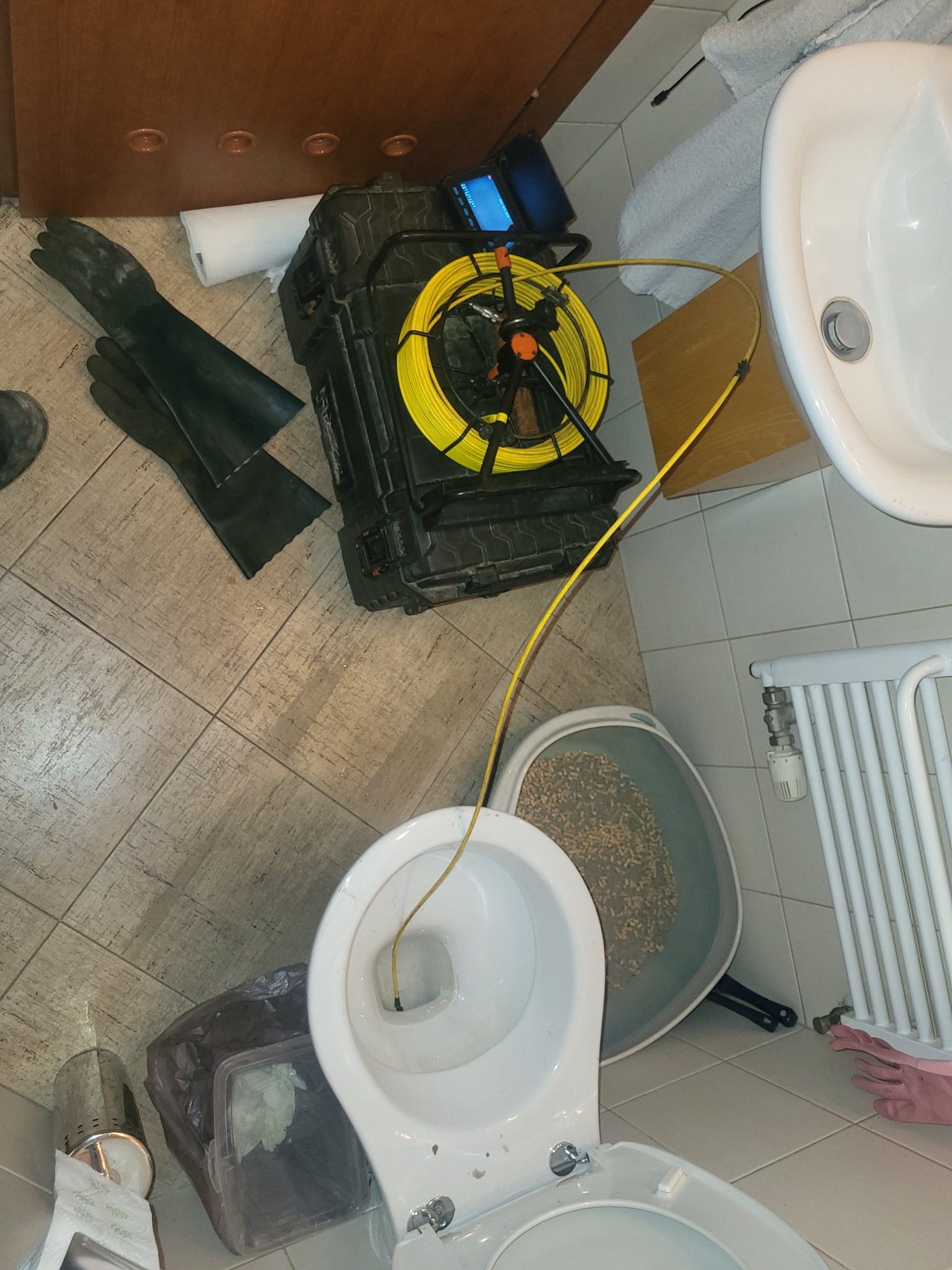 Kamera inspekcyjna z żółtym przewodem w toalecie, obok gumowe rękawice i inne akcesoria. Diagnostyka kanalizacji w trakcie pracy.