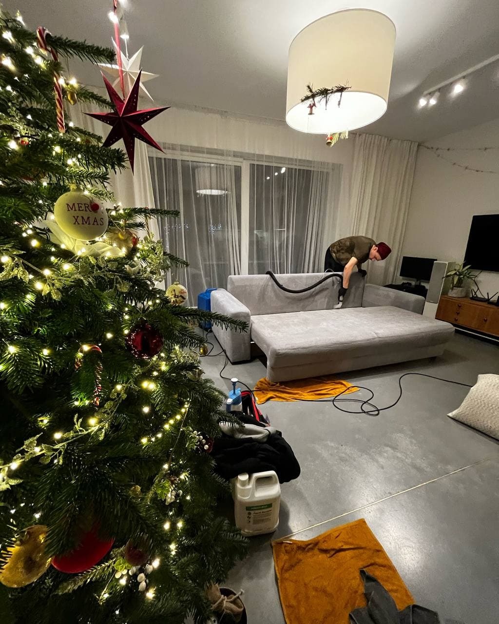 Osoba w czerwonej czapce odkurza szarą kanapę w pokoju z choinką, widoczny sprzęt do czyszczenia i butelka detergentu na podłodze.